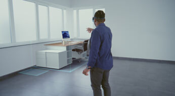 Auf dem symbolischen Bild zum Thema „Was ist Mixed Reality“ steht ein Mann mit Mixed-Reality-Brille in einem leeren Raum und interagiert mit einem digital eingeblendeten Schreibtisch samt Monitor, Stuhl und Kaffeetasse. Die Szene zeigt beispielhaft, wie virtuelle Objekte nahtlos in die reale Umgebung projiziert werden und veranschaulicht das Potenzial von Mixed Reality im Arbeitsalltag.