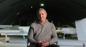 Produktionsstandbild aus der Netflix-Doku „Air Cocaine“: Ein älterer Mann sitzt in einem Flugzeughangar, im Hintergrund stehen zwei kleine Maschinen.