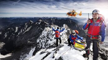 Drei Mitarbeiter:innen der Bergwacht, die sich mit einer geretteten Person auf einem Berggipfel befinden, weisen einen anfliegenden Hubschrauber ein.