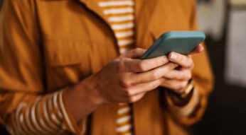 Auf dem Bild zur Nutzung von Meta AI bei WhatsApp ist eine Person zu sehen, die in einem Alltagsszenario ein Smartphone in der Hand hält. Die Person trägt eine gelb-braune Jacke über einem gestreiften Shirt und schaut konzentriert auf das Display. Die Szene vermittelt einen modernen und fokussierten Moment digitaler Kommunikation.