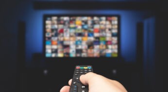 Auf diesem Symbolbild zur Frage „Was kostet Sky 2025“ ist eine Hand mit Fernbedienung zu sehen, die auf einen unscharfen Fernseher mit zahlreichen Streaming-Inhalten gerichtet ist. Das Bild symbolisiert die Vielfalt an Programmen und Paketen, die Sky 2025 bietet, und eignet sich ideal zur Illustration von Kostenübersichten zu Abos und Streaming-Angeboten.