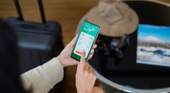 Eine Frau hält ein Smartphone in der Hand und nutzt eine der Reiseplanungs-Apps, um einen Flug von London nach Sydney zu buchen. Im Hintergrund stehen ein schwarzer Koffer, ein Sonnenhut und eine Kamera auf einem runden Tisch, daneben liegt ein Reisemagazin. Die Szene vermittelt Vorfreude und Reiselust.