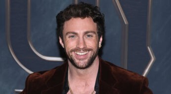 Auf diesem Bild ist Schauspieler Aaron Taylor-Johnson bei einem öffentlichen Auftritt zu sehen. Er trägt ein offenes, schwarzes Hemd unter einem braunen Samt-Sakko und mehrere goldene Ketten. Mit einem breiten Lächeln blickt er direkt in die Kamera. Das Bild lässt sich gut im Kontext aktueller Aaron Taylor-Johnson Filme verwenden.