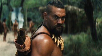 Auf dem Bild aus Chief of War ist Jason Momoa in einer intensiven Szene zu sehen. Er trägt eine Halskette aus Tierzähnen und blickt ernsthaft über die Schulter. Im Hintergrund stehen weitere Stammesmitglieder in traditioneller Kleidung in einer bewaldeten Umgebung. Die Szene wirkt angespannt und eindrucksvoll.