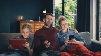 Zum Thema Vodafone GigaTV neue Sender zeigt das Bild eine Familie, die entspannt auf einem grauen Sofa im gemütlichen Wohnzimmer sitzt. Der Vater hält die Fernbedienung und sucht lächelnd nach Sendern, während die Mutter aufs Smartphone blickt. Links vertieft sich der Sohn in sein rotes Tablet. Warmes Licht und eine rote Decke erzeugen eine heimelige, gemütliche Atmosphäre.