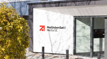 Auf dem Bild zur angekündigten Reality-Show The Hunt ProSieben ist der Eingangsbereich des ProSiebenSat.1 Media SE-Gebäudes bei Tageslicht zu sehen. Das moderne Gebäude zeigt das bekannte rot-schwarze Senderlogo auf weißem Hintergrund. Im Vordergrund befindet sich ein herbstlicher Baum mit gelbem Laub.