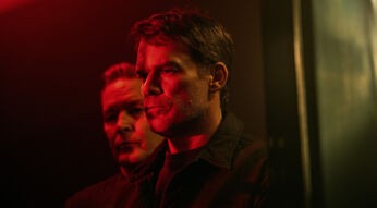 Auf dem Bild zur Serie Dexter Wiedererwachen ist vorne Dexter Morgan, gespielt von Michael C. Hall, mit ernstem Blick zu sehen. Hinter ihm steht Harry Morgan, gespielt von James Remar, im Schatten. Beide wirken angespannt, das rote Licht verstärkt die bedrohliche Atmosphäre.
