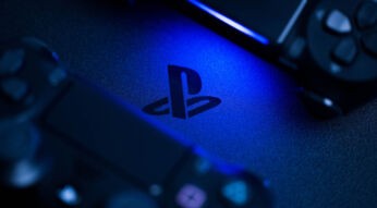 Nahaufnahme des PlayStation-Logos auf einer Konsole, beleuchtet von blauem Licht, mit zwei teilweise sichtbaren Controllern im Vorder- und Hintergrund.