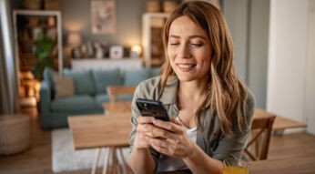 Im Symbolbild für den Handy Garantieverlängerung-Ratgeber sitzt eine lächelnde Frau entspannt in einem modern eingerichteten Wohnzimmer und schaut auf ihr Smartphone.