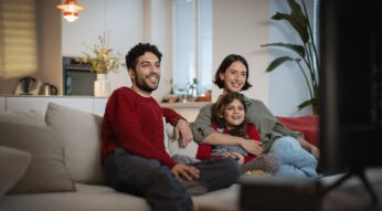 Auf dem Bild zu den internationalen Sendern und TV-Paketen bei Vodafone sitzt eine fröhliche Familie gemeinsam auf dem Sofa und schaut entspannt Fernsehen. Ein Mann, eine Frau und ein Mädchen lachen und wirken völlig in das TV-Programm vertieft.