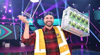 Der Sieger von „Promi Big Brother“ 2025 trägt ein kariertes Hemd, eine gelbe Weste und hält die Siegertrophäe sowie einen Geldkoffer hoch.