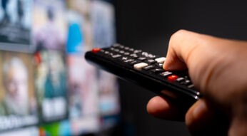 Eine Hand hält eine Fernbedienung und zeigt auf einen TV, der im verschwommen im Hintergrund zu sehen ist.