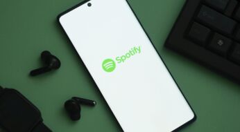 Das Logo von Spotify ist auf einem Handy zu sehen, links davon liegen schwarze Kopfhörer.