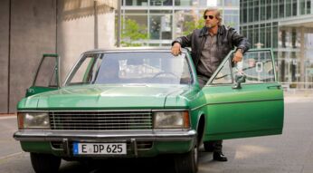Auf dem Bild in den GigaTV-Highlights im Dezember 2025 siehst du eine Szene aus „Der letzte Bulle“: Mick Brisgau (Henning Baum) steht lässig an einem grünen Oldtimer mit offener Tür in einer urbanen Umgebung.