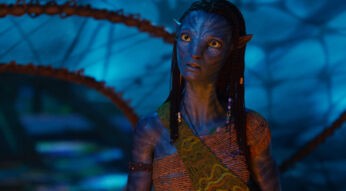 Im Bild aus Avatar: Fire and Ash ist Neytiri, gespielt von Zoe Saldana, in einer nachdenklichen Pose zu sehen. Ihr Gesichtsausdruck ist ernst und wachsam, während sie in einer leuchtend-blauen Umgebung steht. Ihre traditionelle Kleidung und die Zopffrisur unterstreichen ihre kulturelle Identität.