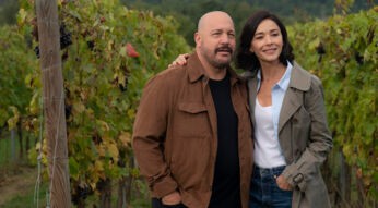 Auf dem Bild zum Film Solo Mio mit Kevin James stehen Matt, gespielt von Kevin James, und Gia, gespielt von Nicole Grimaudo, eng beieinander inmitten eines Weinbergs. Beide wirken gelöst und glücklich – Matt trägt eine braune Jacke, Gia einen beigen Mantel. Die herbstlich gefärbten Reben im Hintergrund erzeugen eine ruhige und romantische Stimmung.