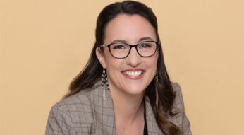 Auf dem Bild ist Abigail Owen, die Autorin der The Games Gods Play-Reihenfolge, vor einem beigefarbenen Hintergrund zu sehen. Sie trägt eine karierte Jacke, eine Brille und auffällige Ohrringe. Mit einem offenen Lächeln wirkt sie sympathisch und professionell – ein stimmiges Autorenportrait für ihre erfolgreiche Fantasyreihe.