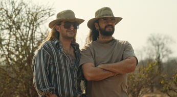 Auf dem Bild zu Kaulitz & Kaulitz Staffel 3 stehen Bill und Tom Kaulitz nebeneinander in einer sonnigen, trockenen Landschaft. Beide tragen breitkrempige Hüte und Sonnenbrillen. Bill ist links in einem gestreiften Hemd zu sehen, Tom rechts mit Bart, schlichtem T-Shirt und verschränkten Armen. Die Szene wirkt entspannt, lässig und vertraut.