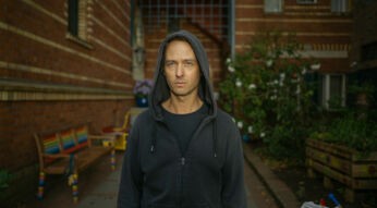 Frontalaufnahme von Tom Schilling in Achtsam Morden. Er trägt einen schwarzen Kapuzenpullover, die Kapuze auf den Kopf.