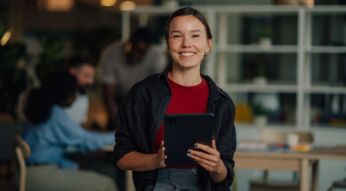 Young businesswoman holding tablet and smiling in modern office with colleagues working in background