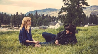 Auf dem Bild zur Dutton Ranch-Serie sitzen Beth Dutton, gespielt von Kelly Reilly, und Rip Wheeler, dargestellt von Cole Hauser, gemeinsam in einer ruhigen Naturlandschaft. Beth sitzt aufrecht im Gras und blickt nachdenklich zu Rip, der entspannt mit Cowboyhut liegt. Die Szene wirkt ruhig, intim und emotional, eingebettet in eine weite, ländliche Kulisse mit Bergen und Bäumen im Hintergrund.
