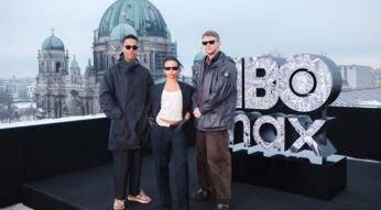 Auf dem Bild zu Industry Staffel 5 stehen die Showrunner Mickey Down und Konrad Kay gemeinsam mit Schauspielerin Myha’la auf einer Dachterrasse in Berlin. Hinter ihnen ist der Berliner Dom zu sehen. Neben der Gruppe steht ein großes HBO-Logo. Alle drei tragen dunkle, moderne Outfits und Sonnenbrillen und blicken selbstbewusst in die Kamera, was eine stylische, promotionale Atmosphäre erzeugt.