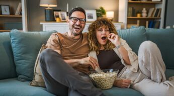 Auf diesem Bild zu gigatv mit netflix sitzt ein entspanntes Paar gemeinsam auf einem Sofa im Wohnzimmer und schaut begeistert Fernsehen. Zwischen ihnen steht eine große Schüssel Popcorn, aus der beide naschen.