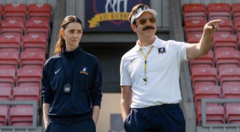 Das Bild zu Neustarts bei Apple TV zeigt eine Szene aus Ted Lasso Staffel 4. Rechts steht Jason Sudeikis als Ted Lasso am Spielfeldrand eines Fußballstadions, mit weißem Poloshirt, Visor und Sonnenbrille, während er selbstbewusst auf das Spielfeld zeigt. Links daneben steht Tanya Reynolds im dunklen Trainingsanzug mit Stoppuhr und beobachtet die Situation mit leicht skeptischem Blick vor roten Stadiontribünen.