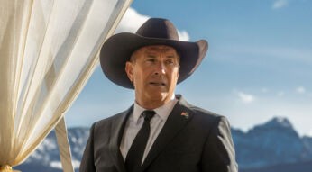 Auf dem Bild zur Yellowstone Serien-Reihenfolge ist John Dutton zu sehen, gespielt von Kevin Costner, der in einem schwarzen Anzug mit Krawatte und Cowboyhut vor einer beeindruckenden Berglandschaft steht. Sein Blick wirkt ernst und nachdenklich, während ein heller Vorhang neben ihm weht. Die Szene vermittelt eine ruhige, aber zugleich würdevolle und leicht angespannte Stimmung.