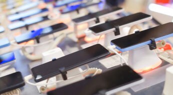 Auf diesem Bild zu Poco oder Redmi sind mehrere moderne Smartphones zu sehen, die in einem Elektronikgeschäft auf Präsentationsständern ausgestellt sind. Die Geräte liegen nebeneinander, sind mit Sicherheitskabeln verbunden und zeigen schwarze Displays. Die Umgebung wirkt hell und technisch, mit unscharfem Hintergrund und farbigen Lichtakzenten, was eine moderne und verkaufsorientierte Atmosphäre vermittelt.