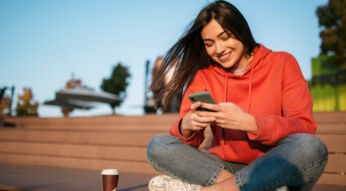Auf dem Bild zum Thema Datenvolumen sparen sitzt eine junge Frau im roten Hoodie entspannt auf einer Holzbank im Freien. Sie lächelt zufrieden, während sie auf ihr Smartphone schaut. Neben ihr steht ein Coffee-to-go-Becher. Die warme Sonne, der blaue Himmel und die lockere Haltung erzeugen eine freundliche, unbeschwerte Stimmung.