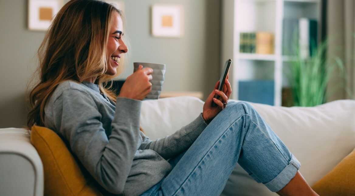 Junge Frau entspannt auf dem Sofa, trink Kaffee und nutzt derweil ihr Smartphone.