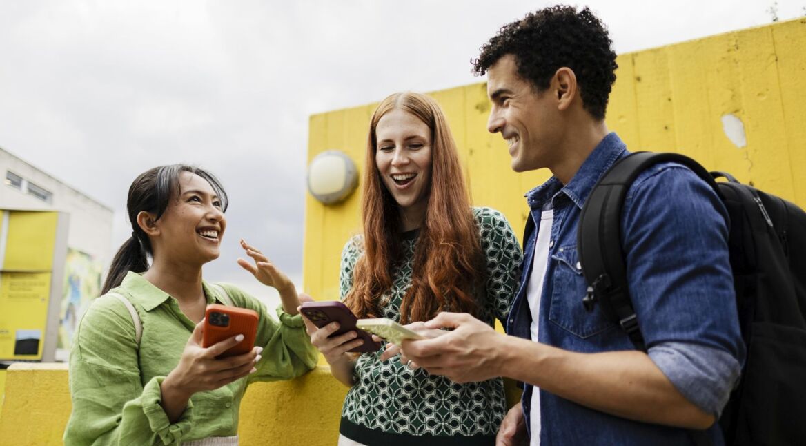 Eine Gruppe Studierende lacht im Freien mit Smartphones in den Händen.