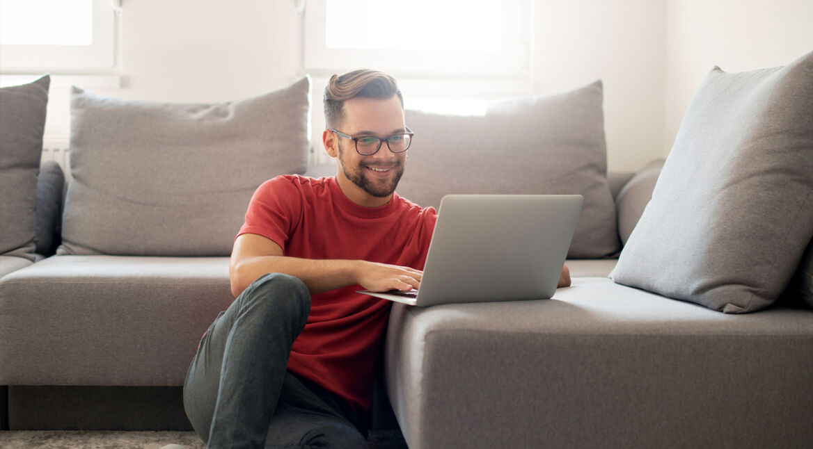 Junger Mann sitzt auf dem Boden vor seinem Sofa und ist lächelnd an seinem Laptop
