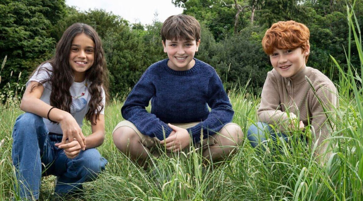 Auf dem Bild zur neuen Harry Potter-Serie sind drei Kinder in einer Wiesenlandschaft zu sehen. In der Mitte sitzt ein Junge mit dunkelblauem Pullover, links von ihm kniet ein Mädchen mit langen dunklen Haaren und weißem T-Shirt, rechts sitzt ein rothaariger Junge in einem beigefarbenen Pullover.