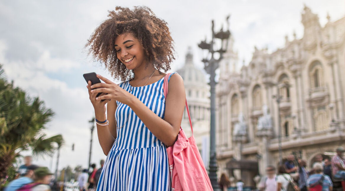 Eine junge Frau steht im Sommerkleid mit blau-weißen Streifen lachend auf einer belebten Straße und schaut auf ihr Smartphone. Im Hintergrund sind prunkvolle historische Gebäude und Palmen zu sehen. Die Szene vermittelt Leichtigkeit und Entdeckerfreude – ideal passend zum Thema Roaming und mobiles Internet auf Reisen.