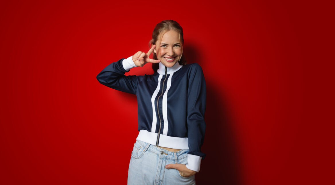 Zum Netflix-Angebot für Vodafone Mobilfunk-Kund:innen zeigt das Bild eine fröhliche junge Frau vor einem kräftig roten Hintergrund. Sie trägt eine sportliche dunkelblaue Jacke mit weißen Streifen und lächelt strahlend, während sie ein Victory-Zeichen mit der rechten Hand macht