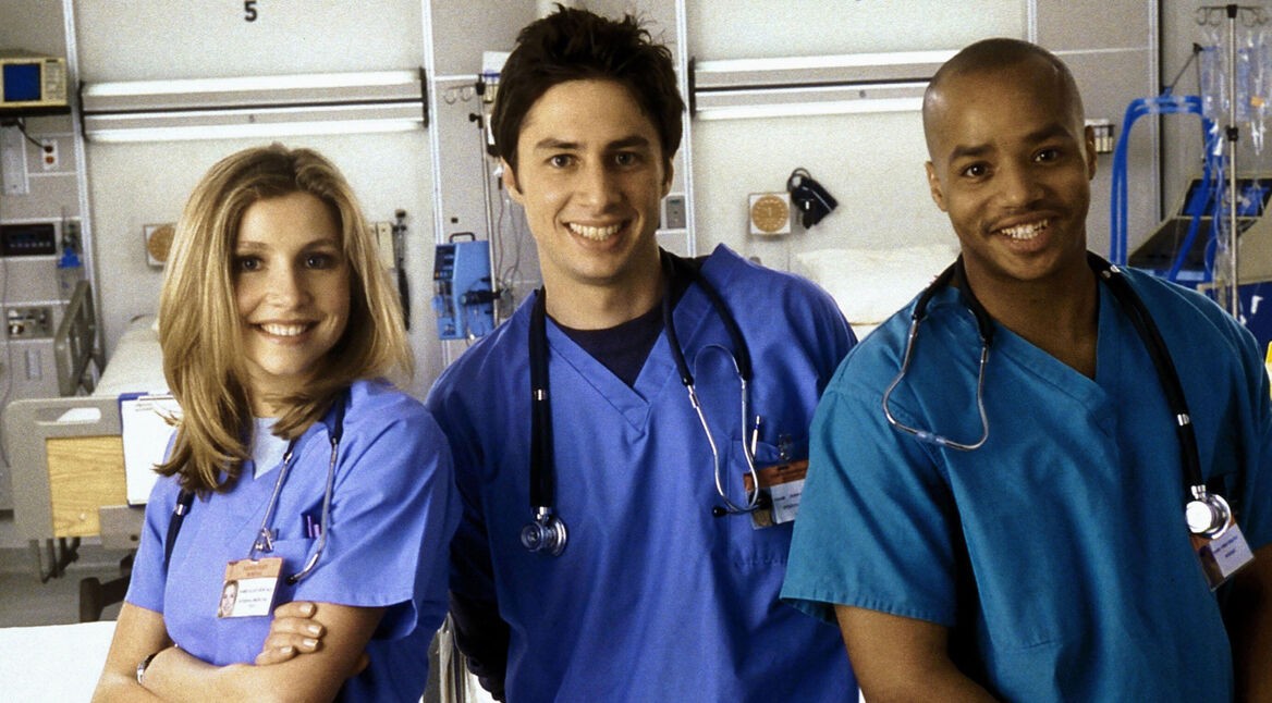Auf dem Bild zu Scrubs beste Folgen sind drei Hauptcharaktere der Serie Scrubs in einem Krankenhaus zu sehen. Links steht Elliot, gespielt von Sarah Chalke, freundlich lächelnd in blauer OP-Kleidung. In der Mitte J.D., dargestellt von Zac Braff, locker und optimistisch. Rechts steht Turk, gespielt von Donald Faison, selbstbewusst und gut gelaunt.
