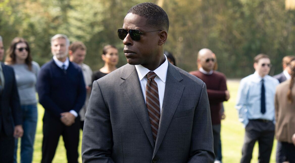 Special Agent Xavier Collins aus „Paradise“ steht im grauen Anzug mit Sonnenbrille auf einer Wiese, mehrere Personen sind unscharf im Hintergrund zu sehen.