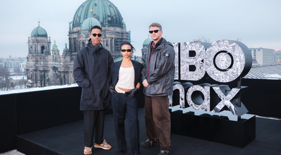 Auf dem Bild zu Industry Staffel 5 stehen die Showrunner Mickey Down und Konrad Kay gemeinsam mit Schauspielerin Myha’la auf einer Dachterrasse in Berlin. Hinter ihnen ist der Berliner Dom zu sehen. Neben der Gruppe steht ein großes HBO-Logo. Alle drei tragen dunkle, moderne Outfits und Sonnenbrillen und blicken selbstbewusst in die Kamera, was eine stylische, promotionale Atmosphäre erzeugt.