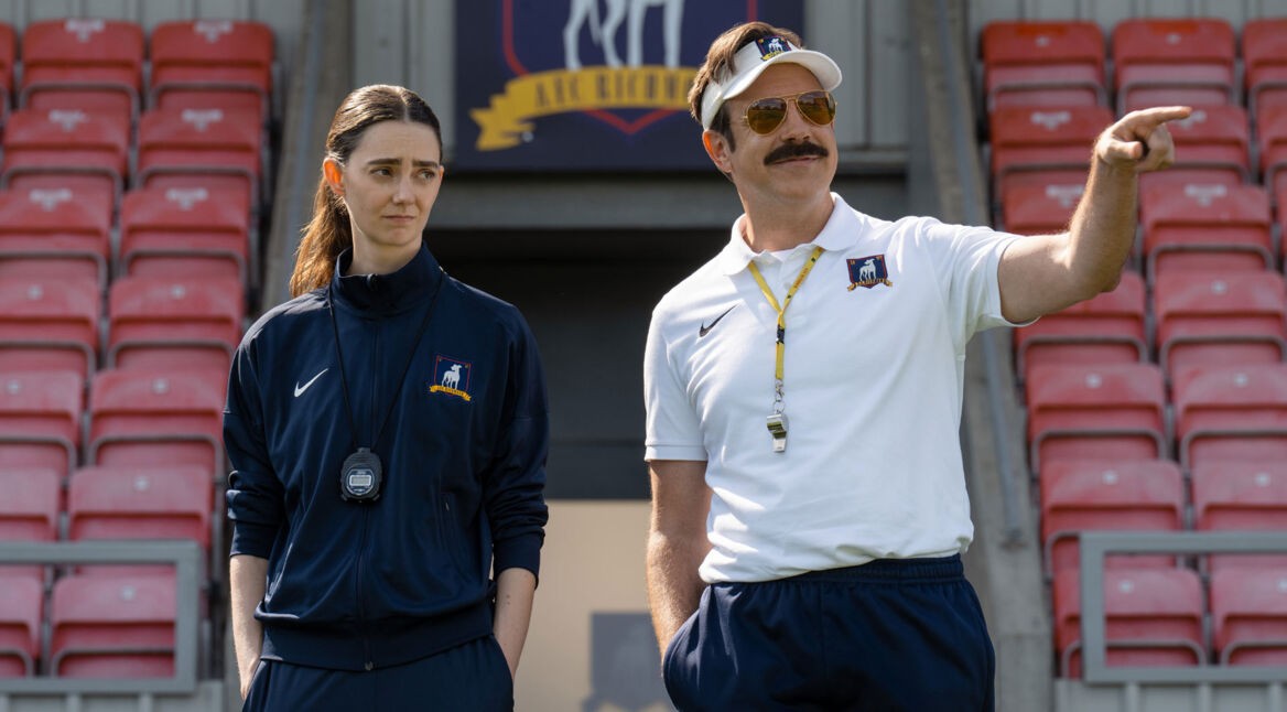 Das Bild zu Neustarts bei Apple TV zeigt eine Szene aus Ted Lasso Staffel 4. Rechts steht Jason Sudeikis als Ted Lasso am Spielfeldrand eines Fußballstadions, mit weißem Poloshirt, Visor und Sonnenbrille, während er selbstbewusst auf das Spielfeld zeigt. Links daneben steht Tanya Reynolds im dunklen Trainingsanzug mit Stoppuhr und beobachtet die Situation mit leicht skeptischem Blick vor roten Stadiontribünen.