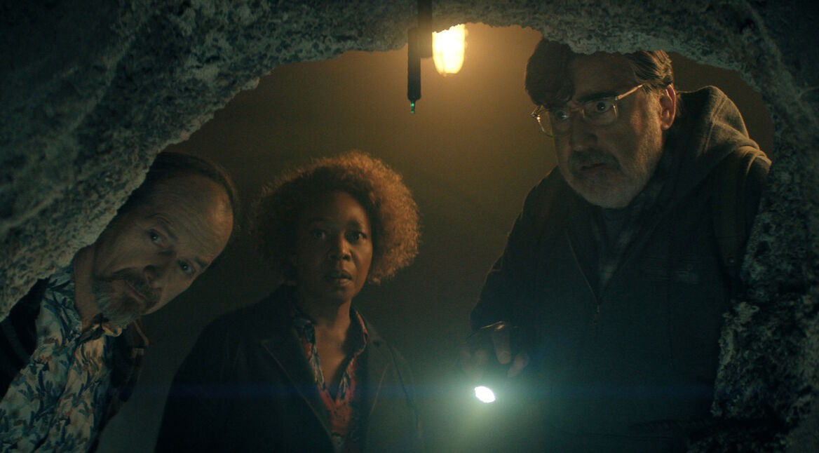 Auf dem Bild zu The Boroughs sind Wally, gespielt von Denis O’Hare, Judy, dargestellt von Alfre Woodard, und Sam, gespielt von Alfred Molina, in einer angespannten Szene zu sehen. Die drei beugen sich durch eine aufgebrochene Betonöffnung und schauen aufmerksam in einen dunklen Raum. Sam hält eine Taschenlampe, deren Licht den Bereich erhellt, während über ihnen eine einzelne Lampe hängt. Ihre ernsten, konzentrierten Blicke verstärken die geheimnisvolle Atmosphäre dieser Szene aus The Boroughs.