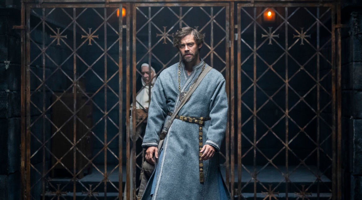 Auf dem Bild zu den GigaTV Highlights im Mai 2026 aus Der Medicus 2 ist Rob Cole, gespielt von Tom Payne, in einer düsteren, mittelalterlichen Szene zu sehen. Er steht im Vordergrund vor einem schweren Gittertor, trägt ein schlichtes graues Gewand mit Gürtel und wirkt erschöpft sowie nachdenklich. Im Hintergrund ist eine weitere Figur schemenhaft erkennbar, was die angespannte und geheimnisvolle Stimmung unterstreicht.