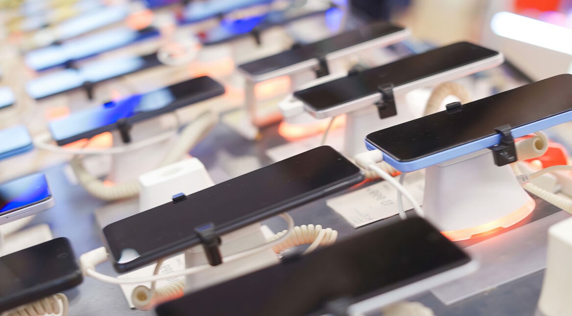 Auf diesem Bild zu Poco oder Redmi sind mehrere moderne Smartphones zu sehen, die in einem Elektronikgeschäft auf Präsentationsständern ausgestellt sind. Die Geräte liegen nebeneinander, sind mit Sicherheitskabeln verbunden und zeigen schwarze Displays. Die Umgebung wirkt hell und technisch, mit unscharfem Hintergrund und farbigen Lichtakzenten, was eine moderne und verkaufsorientierte Atmosphäre vermittelt.