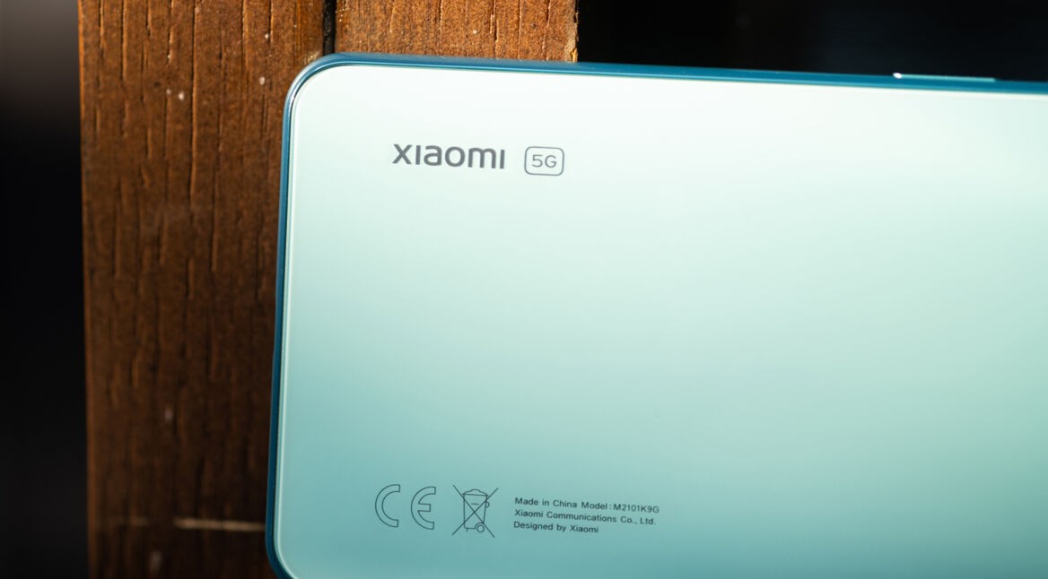 Auf dem Bild zum Xiaomi 5G Handy ist die Rückseite eines modernen Smartphones in einem hellen Mintgrün zu sehen. Oben links befindet sich das Xiaomi-Logo mit 5G-Kennzeichnung, während unten technische Angaben und Zertifizierungssymbole abgebildet sind. Das Gerät liegt vor einer warmen Holzoberfläche und vermittelt eine hochwertige, minimalistische Produktstimmung.