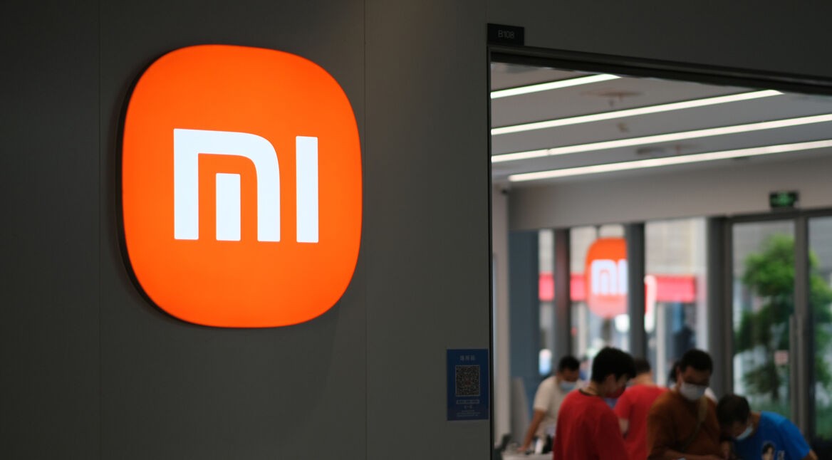 Auf dem Bild mit dem Fokus auf Xiaomi Modelle Reihenfolge ist das leuchtend orangefarbene Xiaomi-Logo gut sichtbar an einer Innenwand eines modernen Stores angebracht. Im Hintergrund erkennt man einen hellen Verkaufsraum mit mehreren Kunden, die sich Produkte ansehen. Die Szene wirkt ruhig, modern und vermittelt eine technikorientierte Einkaufsatmosphäre.