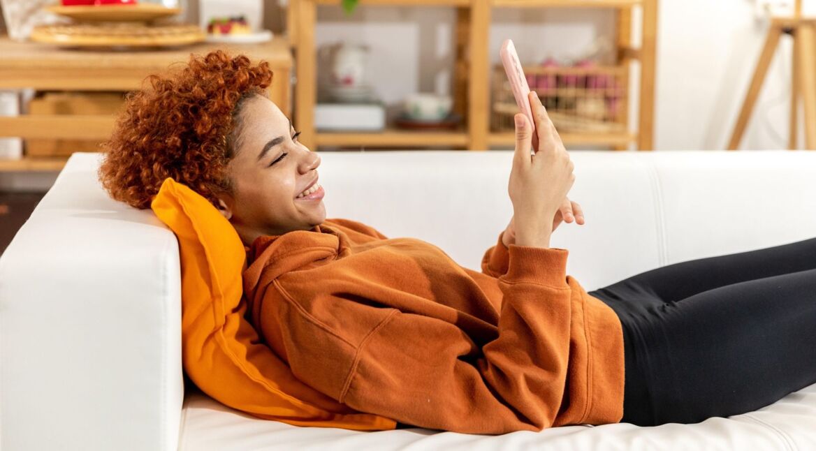 Schwarze Frau sitzt auf der Couch mit ihrem Smartphone und lächelt es an.