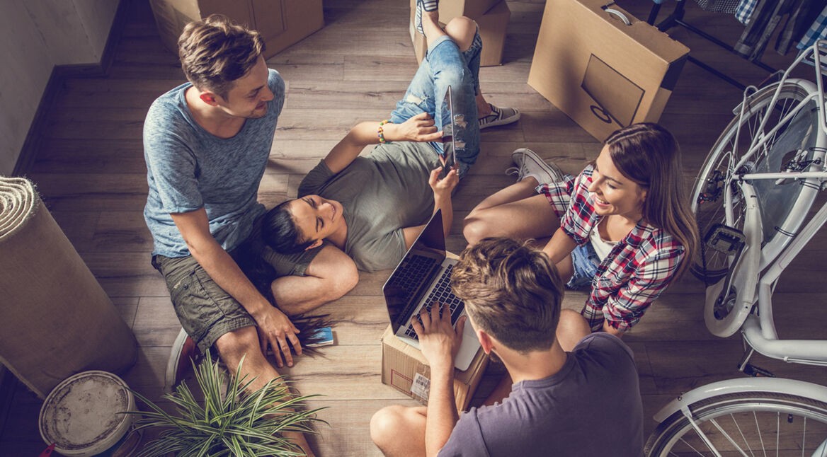 Vier junge Leute, die sich über WG-Portale gefunden haben, sitzen auf dem Boden zwischen Umzugskartons