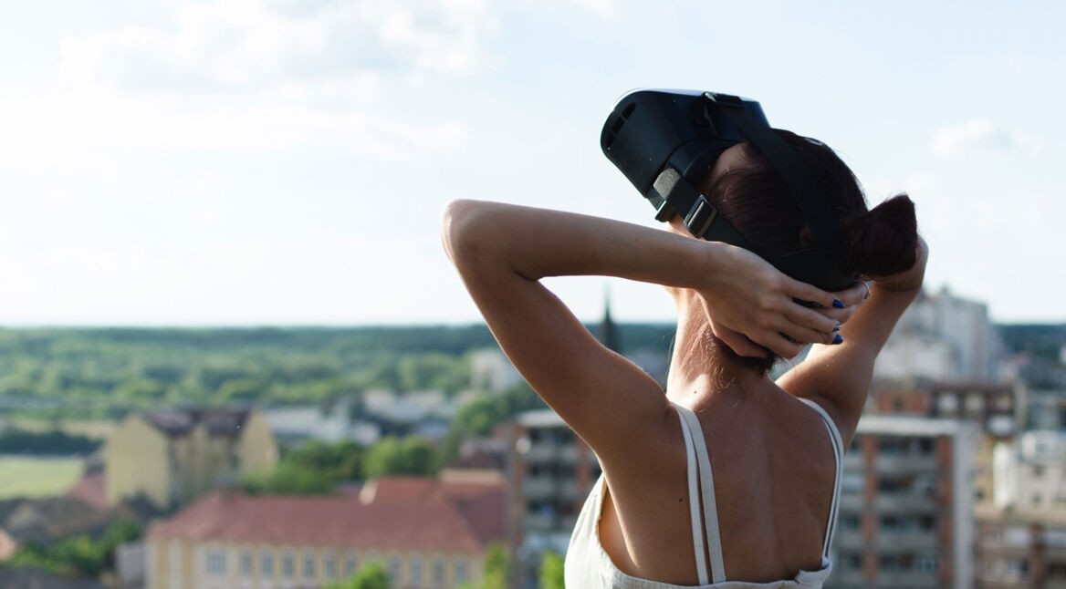 Eine Frau mit VR-Headset steht auf dem Dach und legt die Arme in den Kopf, im Hintergrund sieht man Häuser und Dächer einer Stadt.