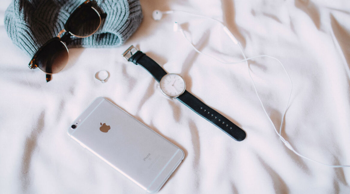 iPhone liegt neben Mütze, Sonnenbrille und Uhr auf einem Bett.